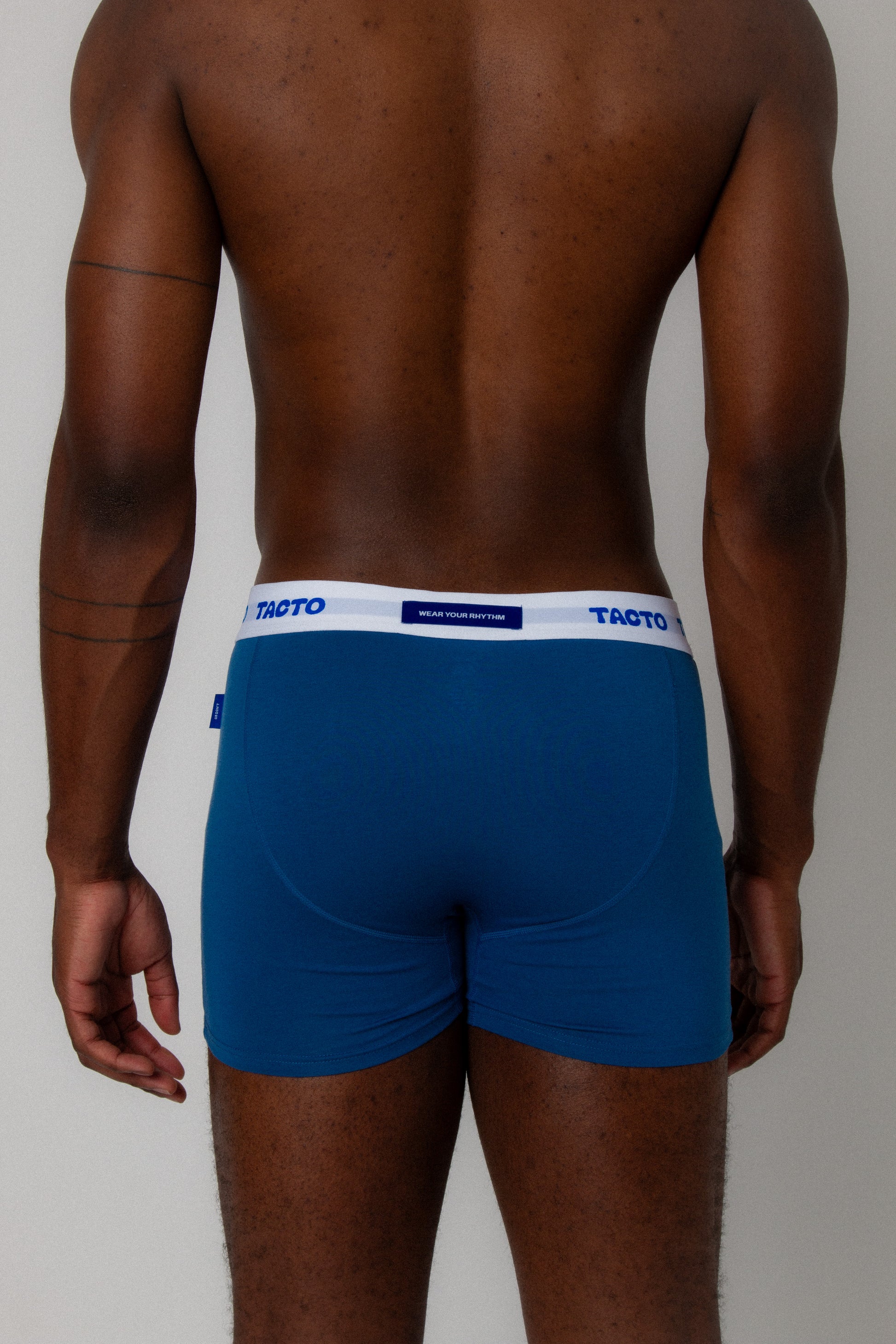 Person wearing blue boxer briefs with a visible brand logo on a plain background. TACTO Collective underwear. . WEAR YOUR RHYTHM blue back tag