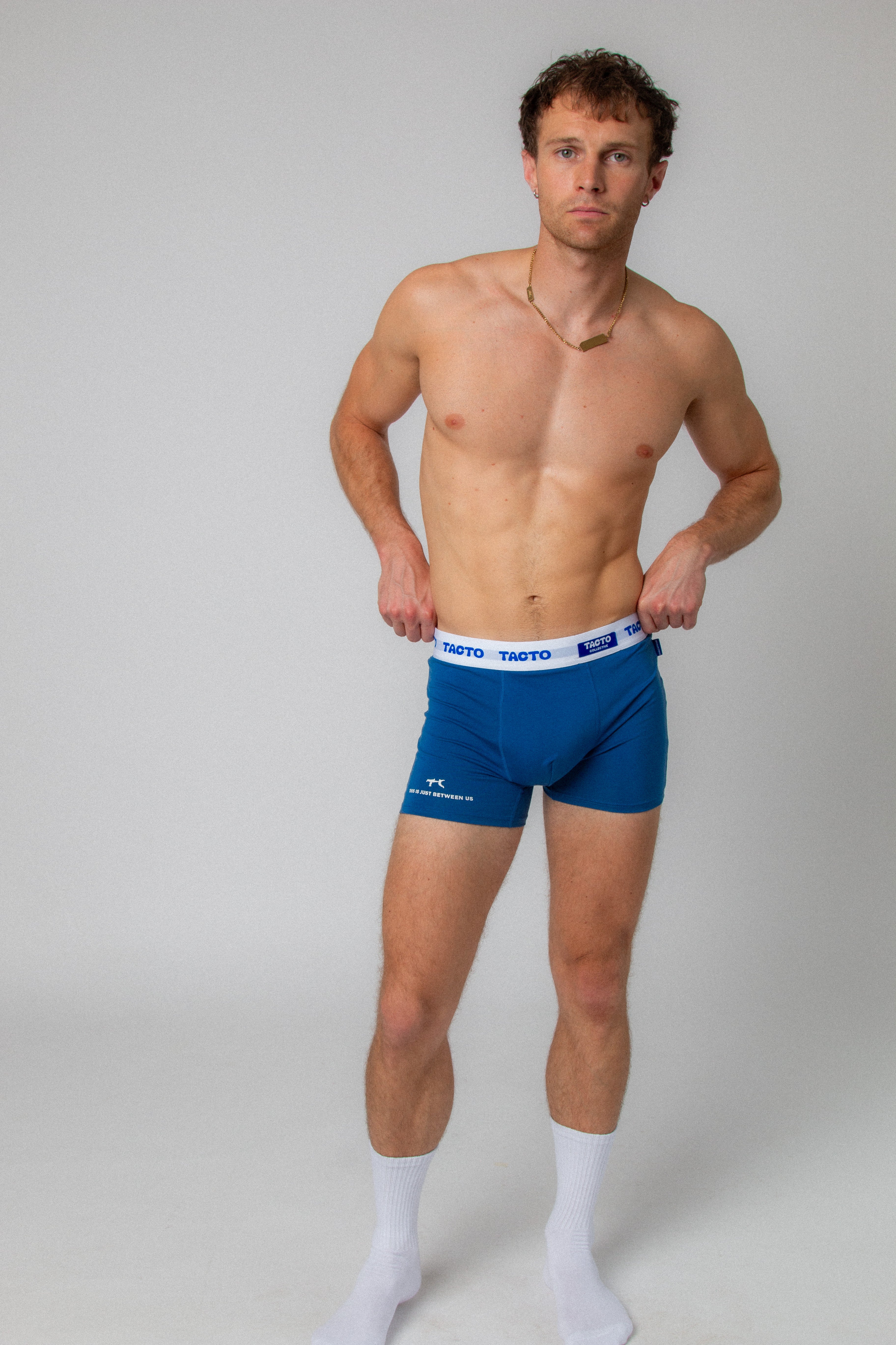 Man wearing blue boxer briefs with a white waistband on a gray background. TACTO Collective underwear.