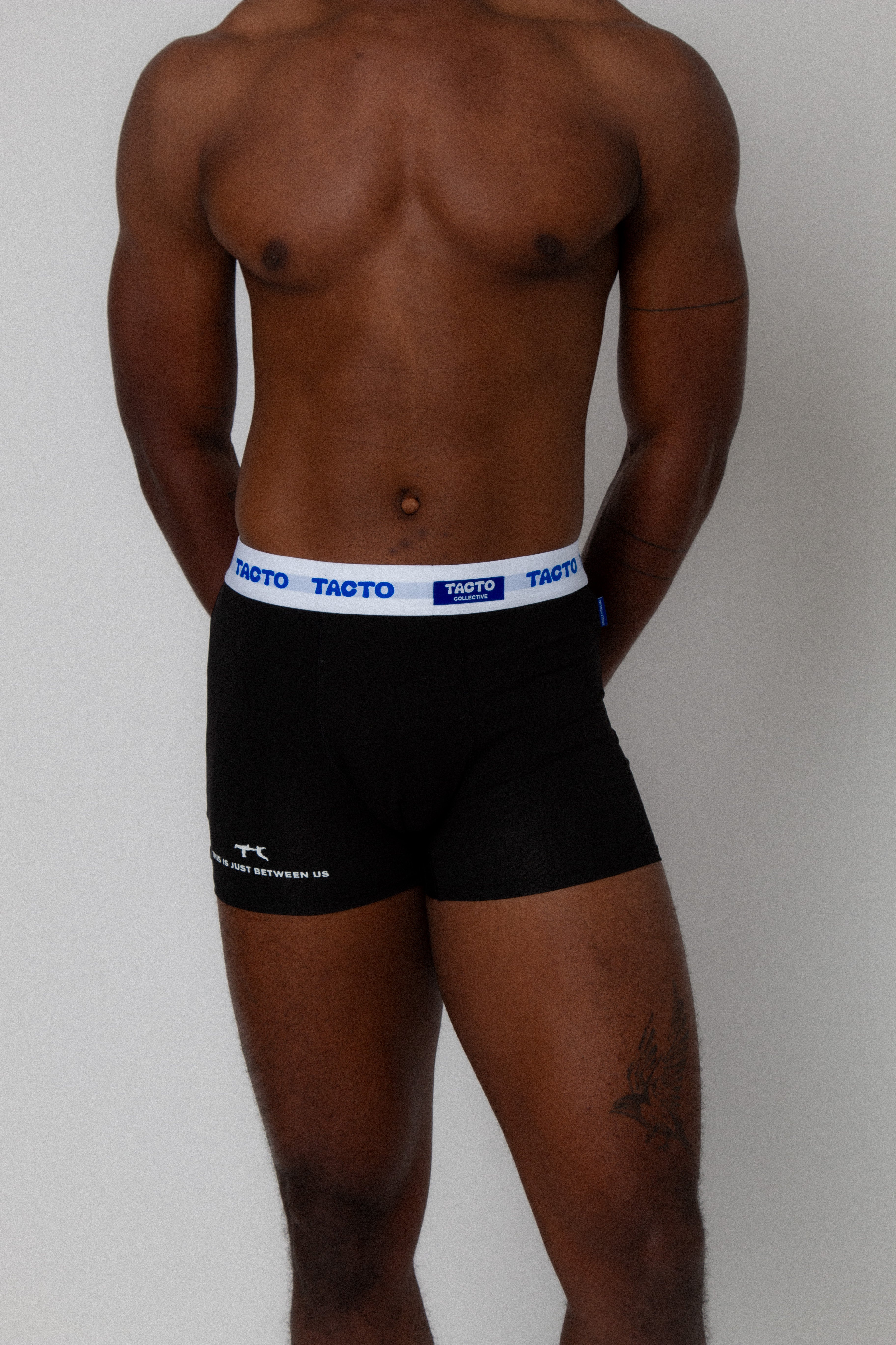 Person wearing black boxer briefs with a visible brand logo TACTO on a plain background. Detail on leg THIS IS JUST BETWEEN US