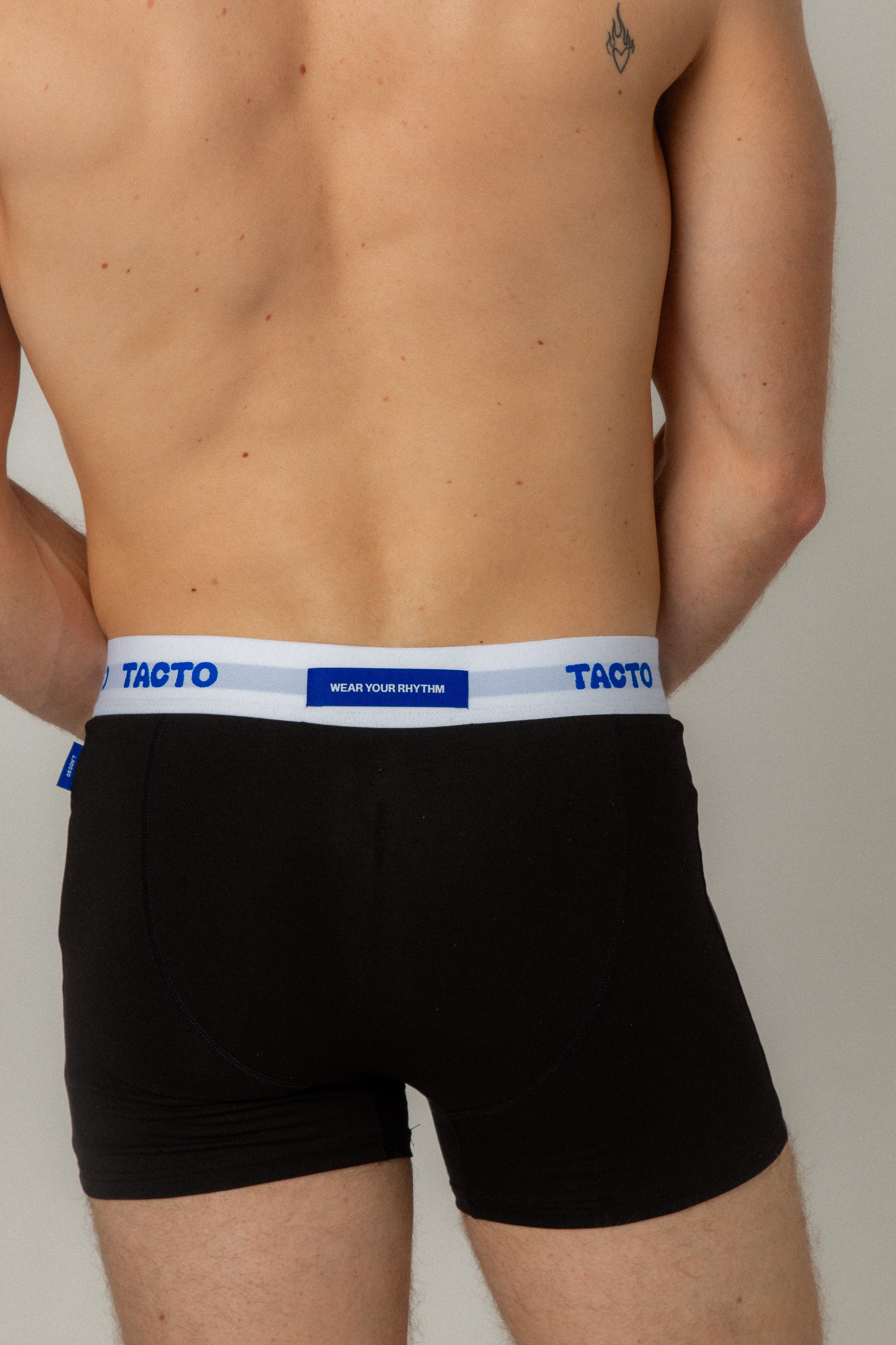 Black boxer briefs with white waistband featuring 'Tacto' branding on a plain background. Wear Your Rhythm. Blue tags