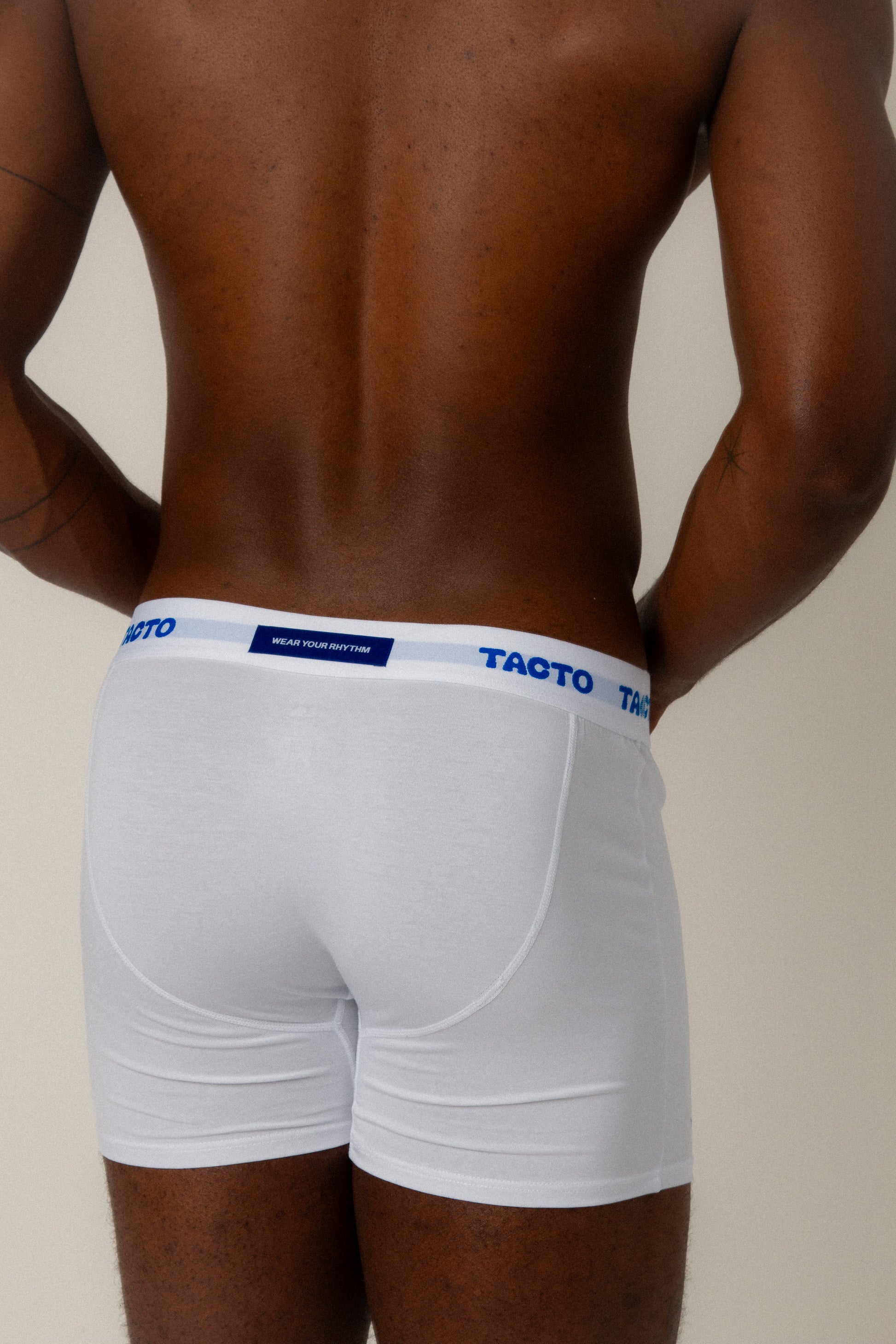 Person wearing white boxer briefs with blue waistband on a plain background. Blue back tag that read WEAR YOUR RHYTHM