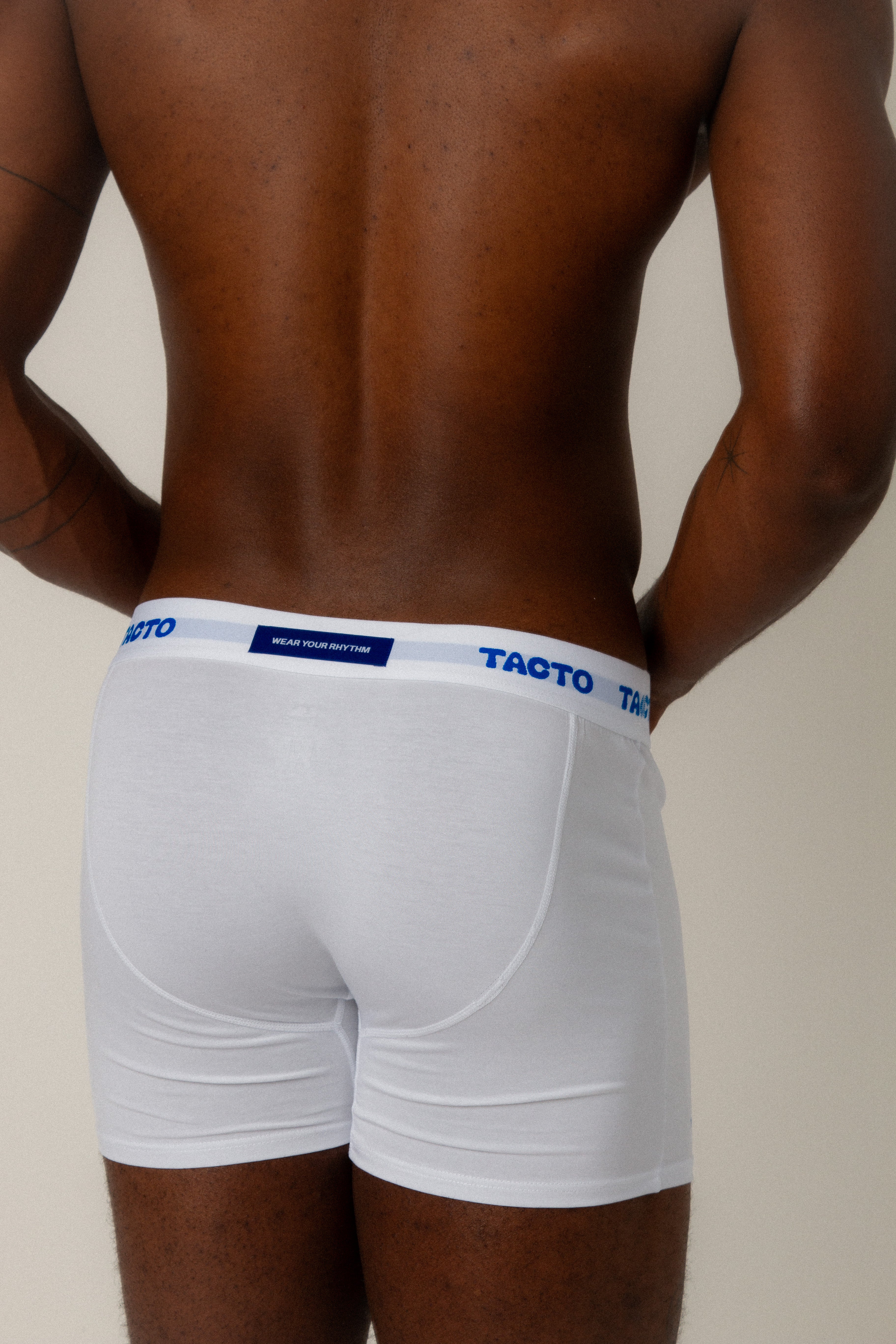 Person wearing white boxer briefs with blue waistband on a plain background. Blue back tag that read WEAR YOUR RHYTHM